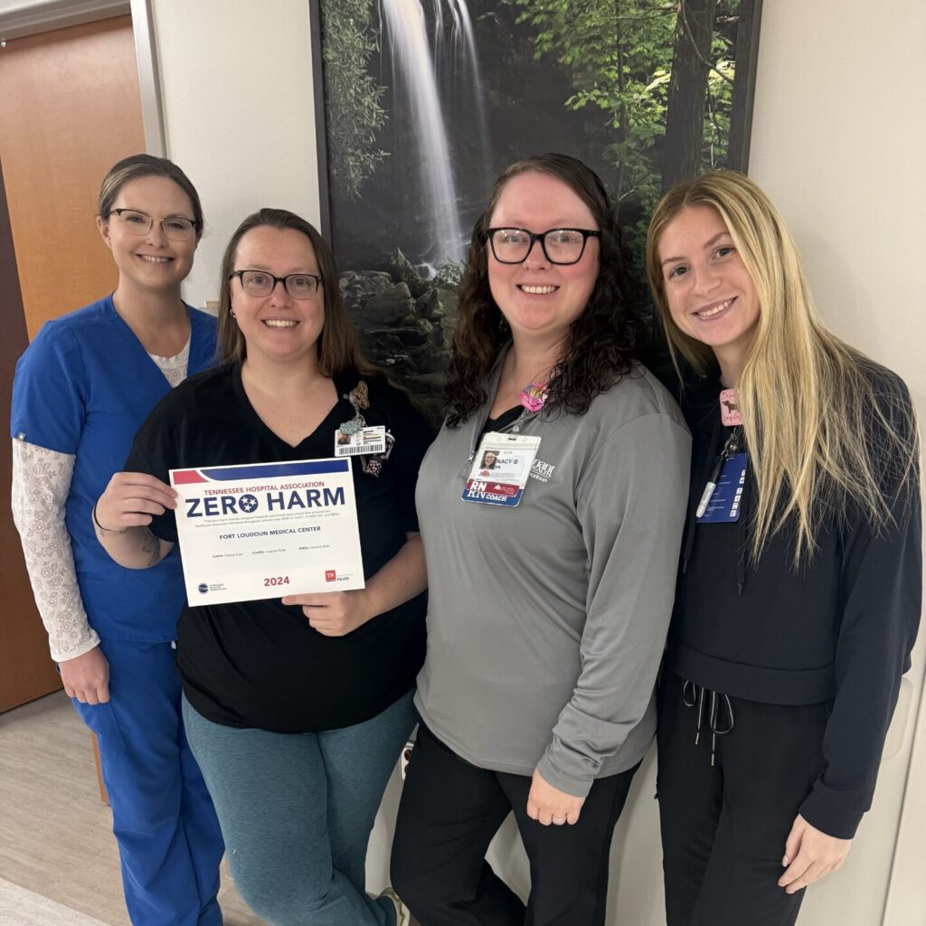 Four women stand indoors smiling at the camera. One woman holds a Zero Harm certificate for 2024. They are dressed in work attire, standing in front of a nature-themed wall art.