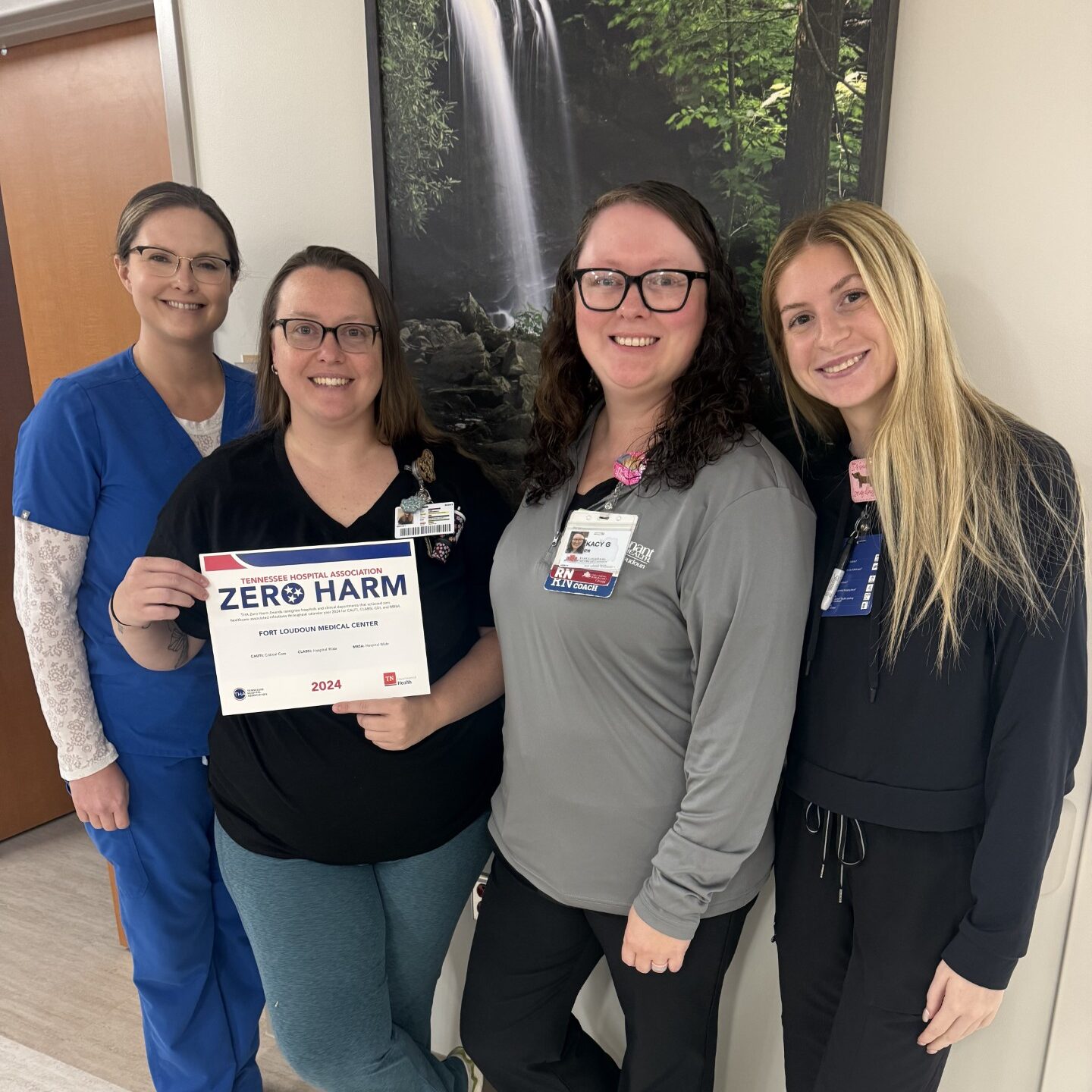 Four women stand indoors smiling at the camera. One woman holds a Zero Harm certificate for 2024. They are dressed in work attire, standing in front of a nature-themed wall art.