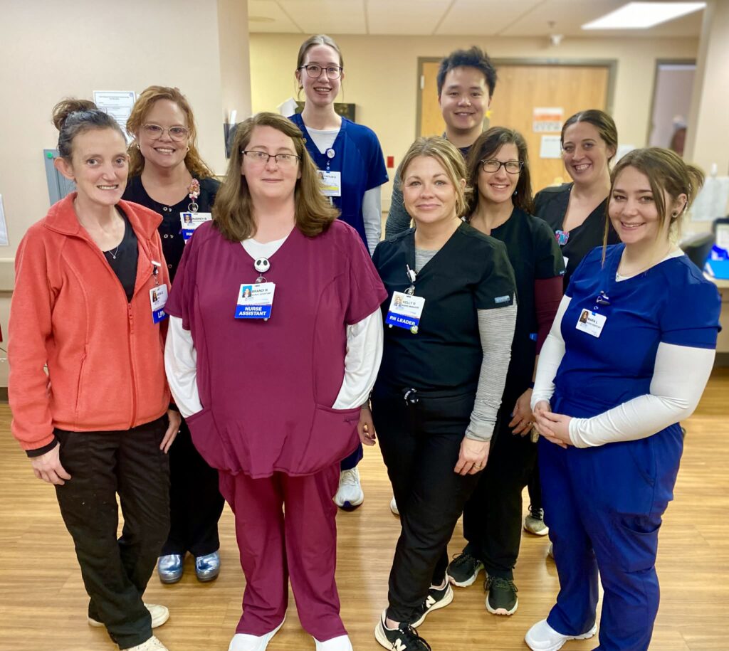 A group of nine smiling healthcare workers, wearing scrubs and name badges, stand together in a brightly lit Methodist Medical Center hallway with wood flooring.