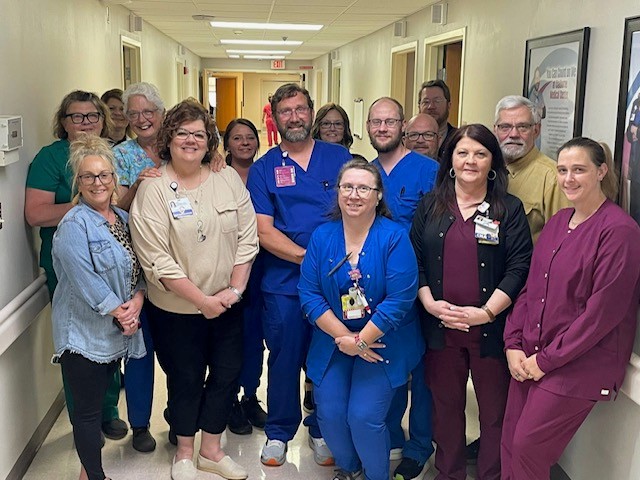 A group of healthcare professionals in scrubs and office attire stand smiling together in a hospital hallway, posing for a group photo under bright lights.
