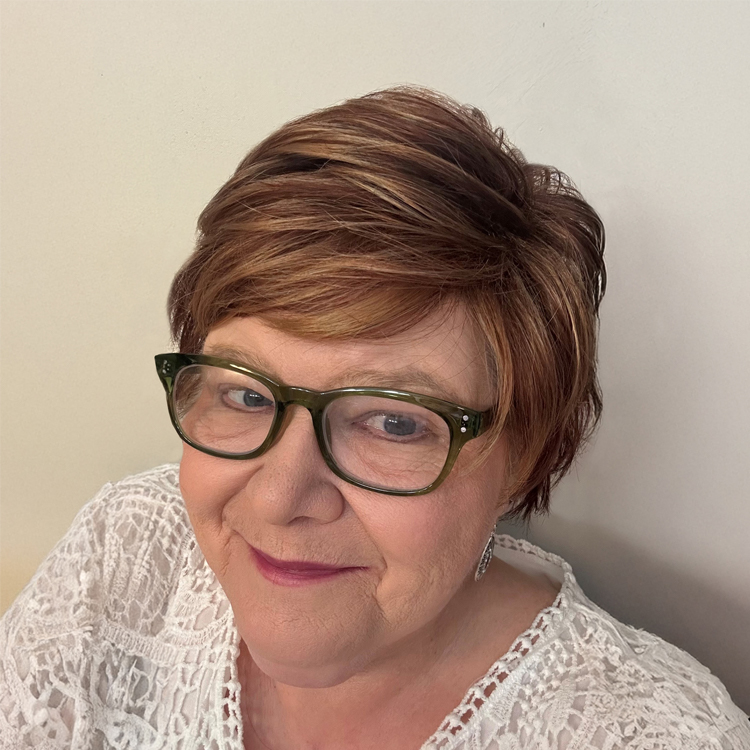 A woman with short, layered brown hair and green glasses smiles gently. She is wearing a white lace top and silver earrings, posing in front of a plain light-colored wall.