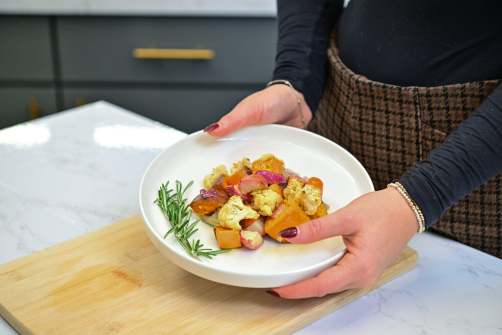 A person holding a white plate with roasted vegetables and bread pieces, garnished with a sprig of rosemary, above a wooden cutting board on a white countertop.
