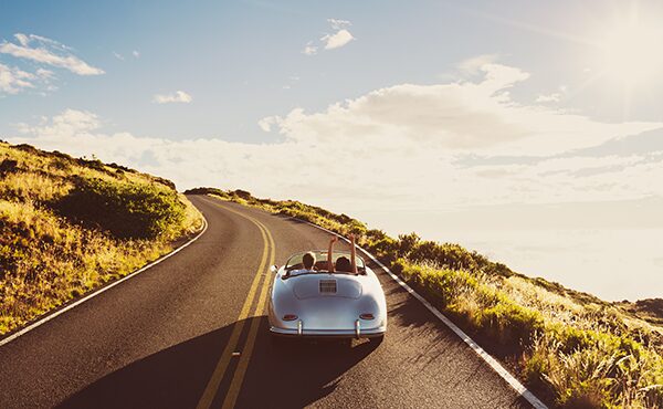 A silver convertible drives along a winding road surrounded by grassy hills under a bright, sunny sky. Two people sit inside, and one raises an arm in excitement as they head toward the horizon.