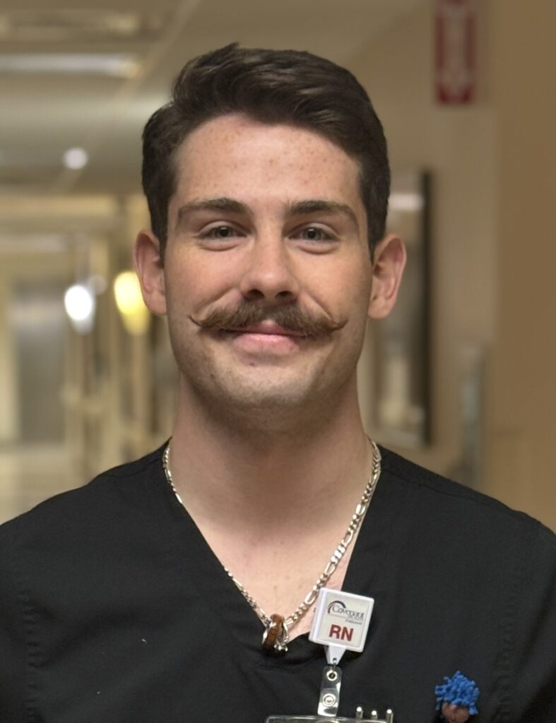 A male nurse with dark hair and a styled mustache smiles at the camera. He is wearing black scrubs, a necklace, and a badge that says RN in a hospital hallway, proudly celebrating as Matt Pack wins DAISY Award.