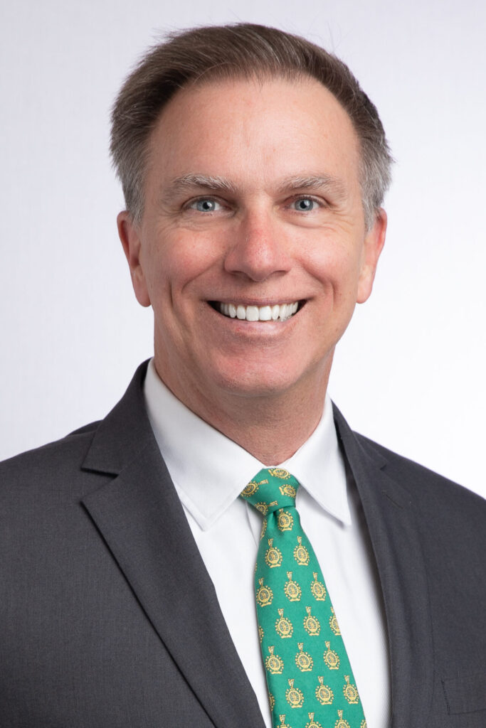 A man in a dark suit, white shirt, and green patterned tie smiles in a professional headshot against a plain light background.