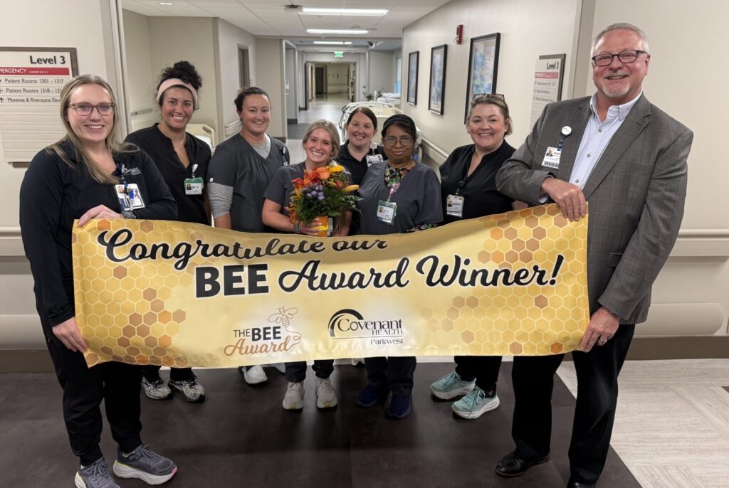A group of eight smiling healthcare workers stand in a hospital hallway, holding a banner that reads Congratulations Madison Taggart BEE Award Winner! One person in the center holds a bouquet of flowers.