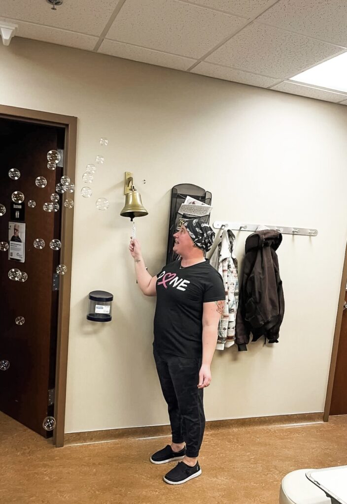 A person in casual clothes and a headscarf smiles while ringing a golden bell in a breast medical office, as bubbles float nearby. Coats hang on hooks and a white exam table is visible in the room.