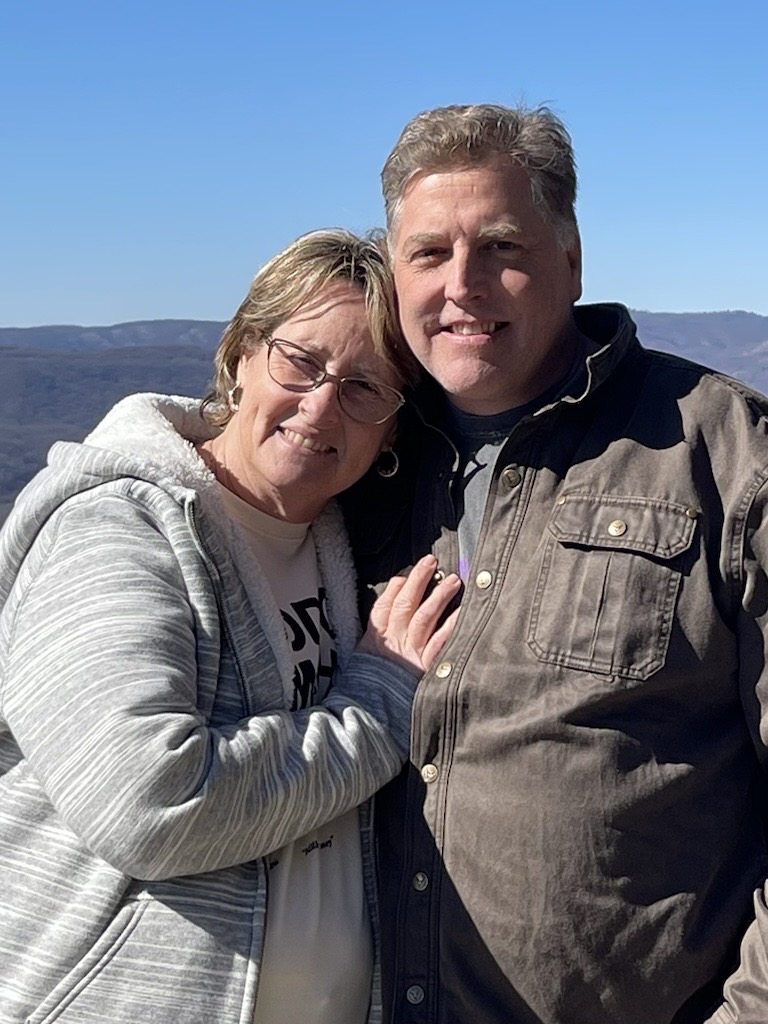 A smiling couple stands close together outdoors, with mountains and a clear blue sky in the background. The woman wears glasses and a striped jacket, while the man wears a brown jacket.