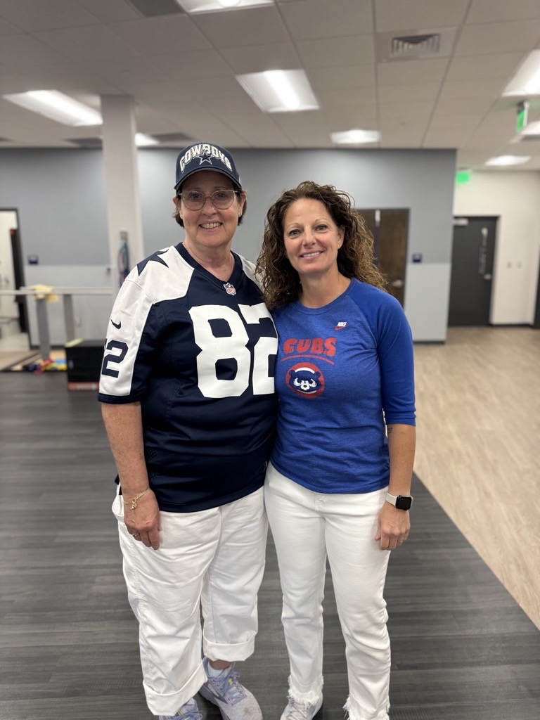 Two women stand together indoors, smiling. One wears a Dallas Cowboys jersey and cap with white pants; the other wears a Chicago Cubs t-shirt with white pants. The background shows an office setting with desks and doors.