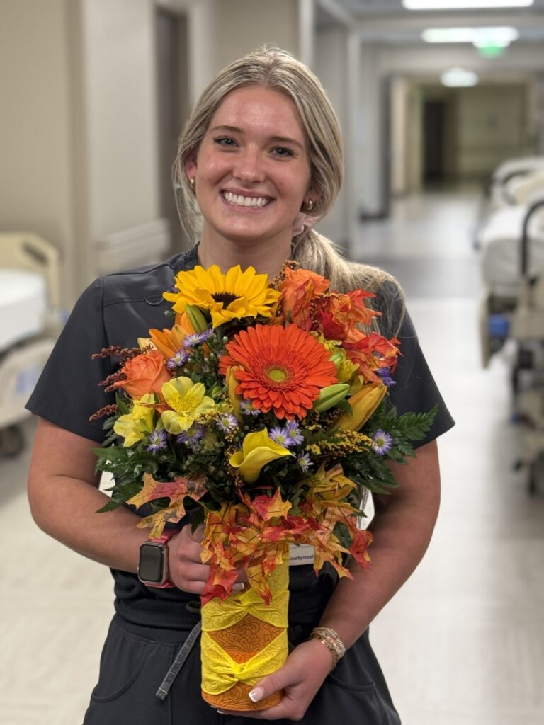 A smiling woman in scrubs stands in a hospital hallway, proudly holding a colorful bouquet after receiving the Madison Taggart BEE Award. Hospital beds and equipment are visible in the background.