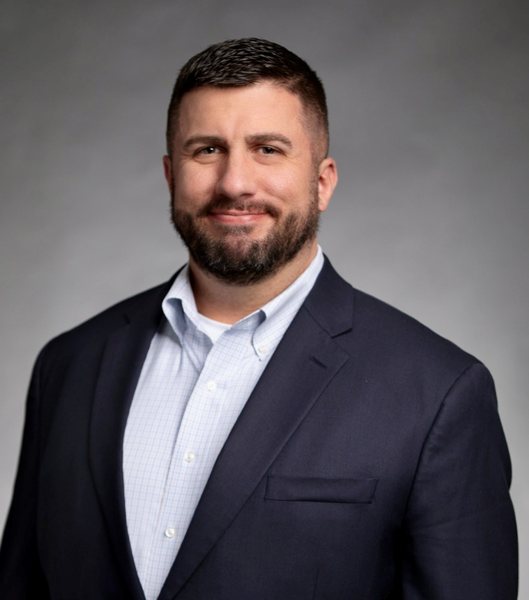 A man with short brown hair and a beard, wearing a navy blue suit jacket over a light blue checkered shirt, smiles at the camera against a plain gray background, embodying professionalism ideal for nursing jobs at Covenant Health.