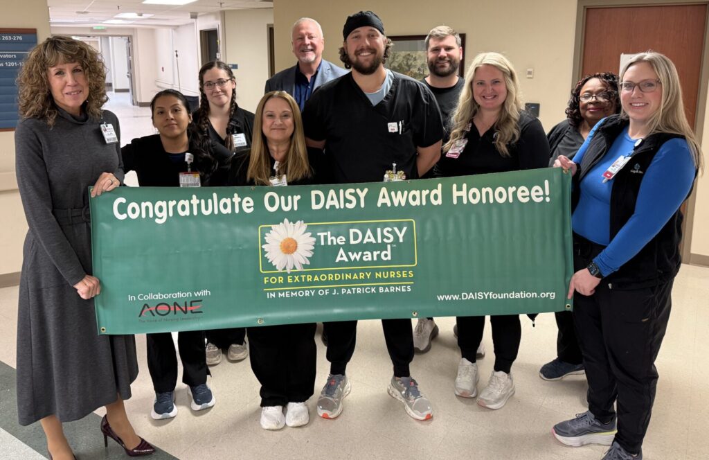A group of nurses and staff stand indoors, smiling and posing with a green banner that reads, "Congratulate Our DAISY Award Honoree!" as Larissa Iurco wins the DAISY Award for Extraordinary Nurses.
