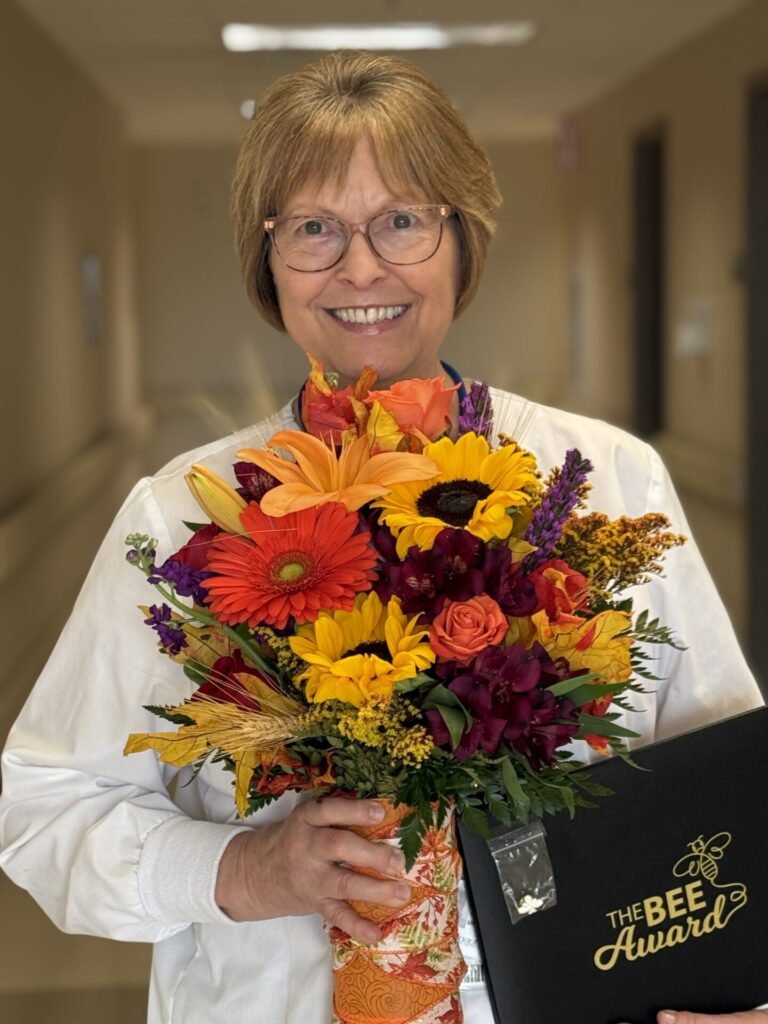 A smiling woman in glasses and a white jacket holds a colorful bouquet of flowers and a black folder that says Madison Taggart BEE Award in a hallway.