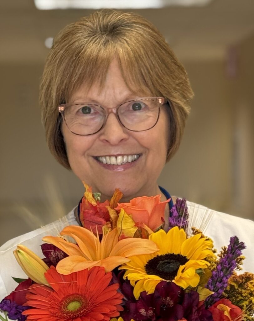 A smiling woman with short light brown hair and glasses, Pam Reuland BEE Award recipient, holds a colorful bouquet of flowers—orange lilies, yellow sunflowers, and red daisies—indoors with a softly blurred background.