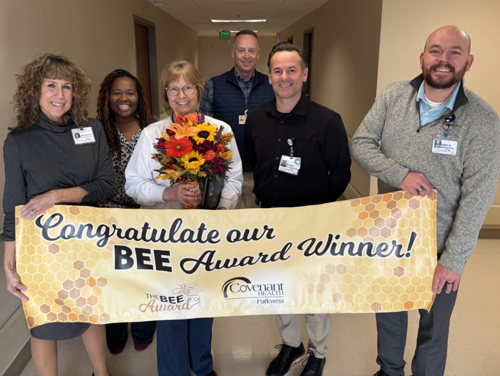 Six smiling people stand in a hallway. The woman in the center, Madison Taggart, holds a bouquet of flowers. They hold a banner with a honeycomb pattern reading &ldquo;Congratulate our BEE Award Winner!&rdquo;.