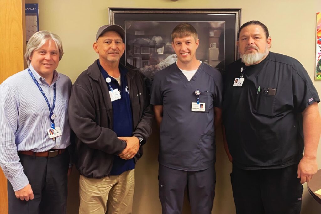 Four men stand side by side indoors, smiling at the camera. Three wear work uniforms or scrubs with ID badges, and one wears business attire. A framed picture hangs on the wall behind them.