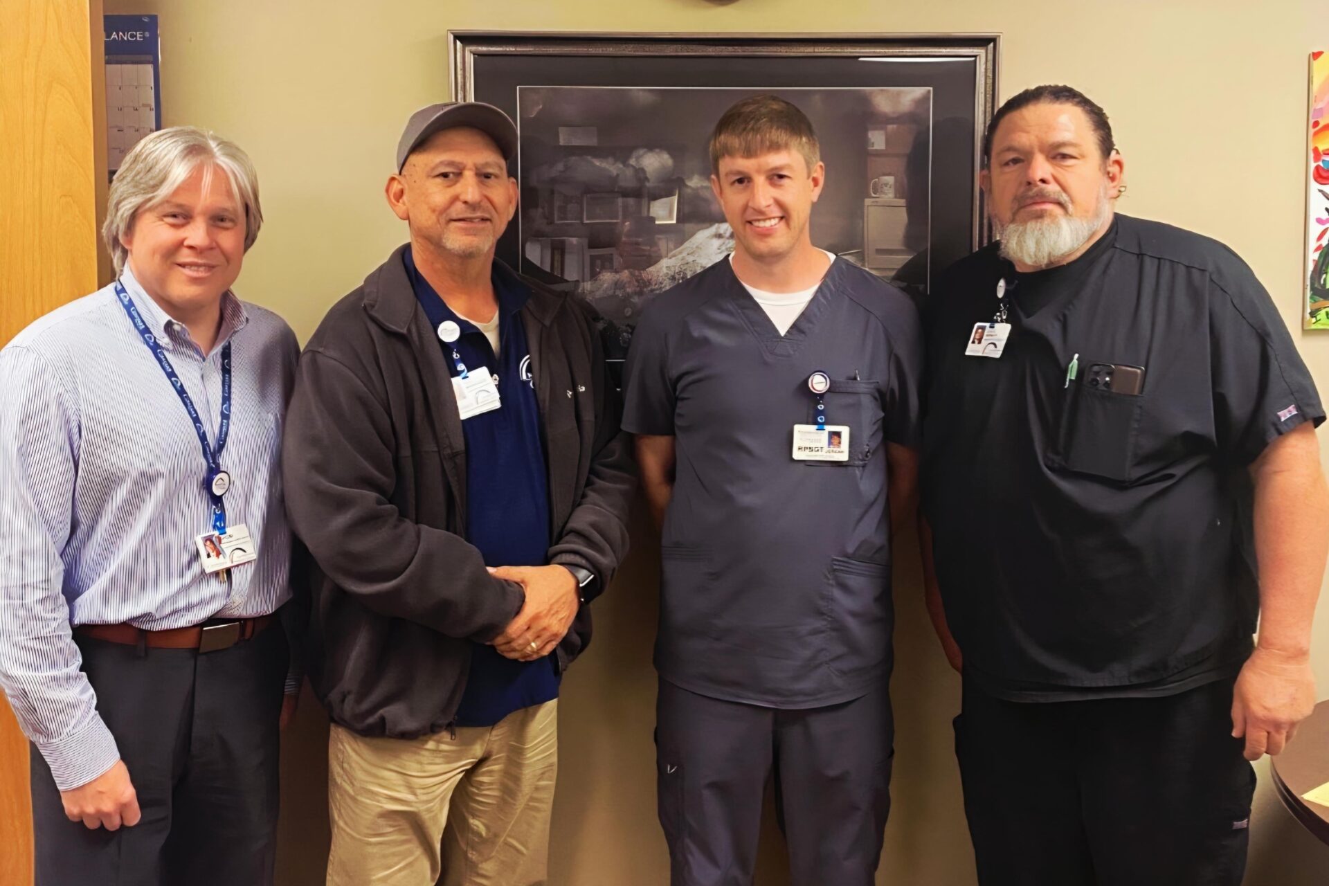 Four men stand side by side indoors, smiling at the camera. Three wear work uniforms or scrubs with ID badges, and one wears business attire. A framed picture hangs on the wall behind them.
