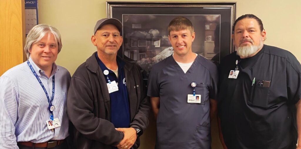 Four men stand side by side indoors, smiling for a photo. Three wear dark medical scrubs with ID badges, while one wears a light button-down shirt and tie. They gather to celebrate an accreditation milestone, with a framed picture visible on the wall behind them.