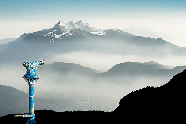 A blue coin-operated binocular viewer overlooks misty mountain valleys and distant snow-capped peaks under a clear sky. The landscape is layered with dark silhouettes and light fog.