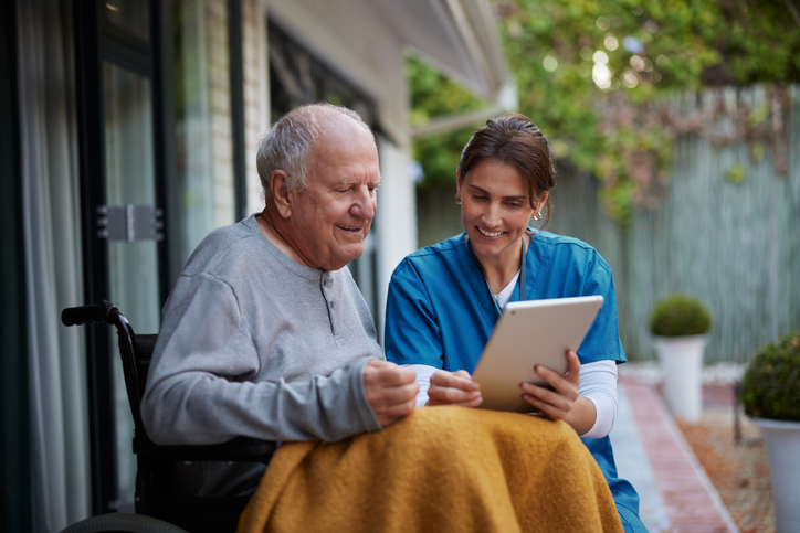 A nurse in blue scrubs sits beside an elderly man in a wheelchair, showing him a tablet.