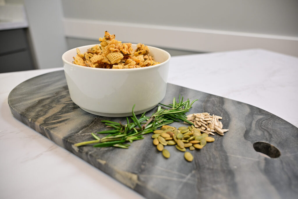 A white bowl of chex mix sits on a gray marble board with fresh rosemary, pumpkin seeds, and sunflower seeds arranged beside it on a white countertop.