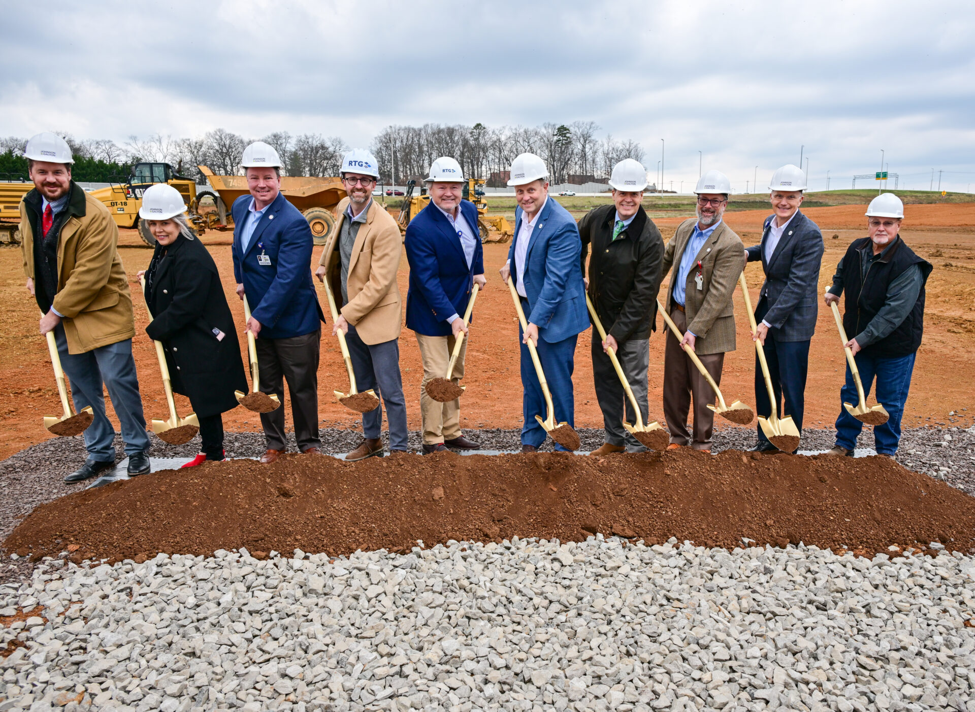 Ten people in hard hats hold shovels and pose for a groundbreaking ceremony at a construction site.