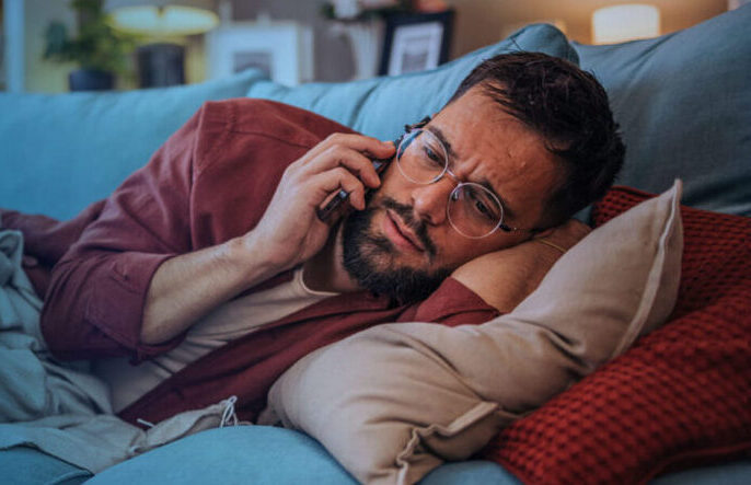 A man lies on a couch, looking under the weather and talking on a smartphone.