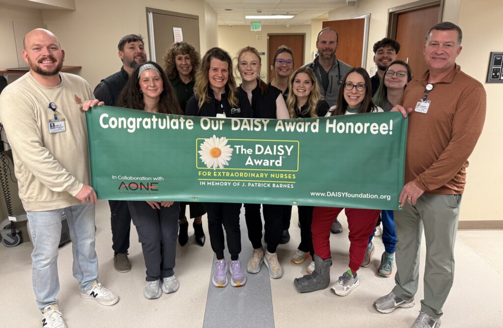 A group of smiling healthcare workers stands in a hallway holding a green banner that reads, &ldquo;Congratulate Our DAISY Award Honoree! Ethan Howe wins DAISY Award for Extraordinary Nurses.&rdquo;.