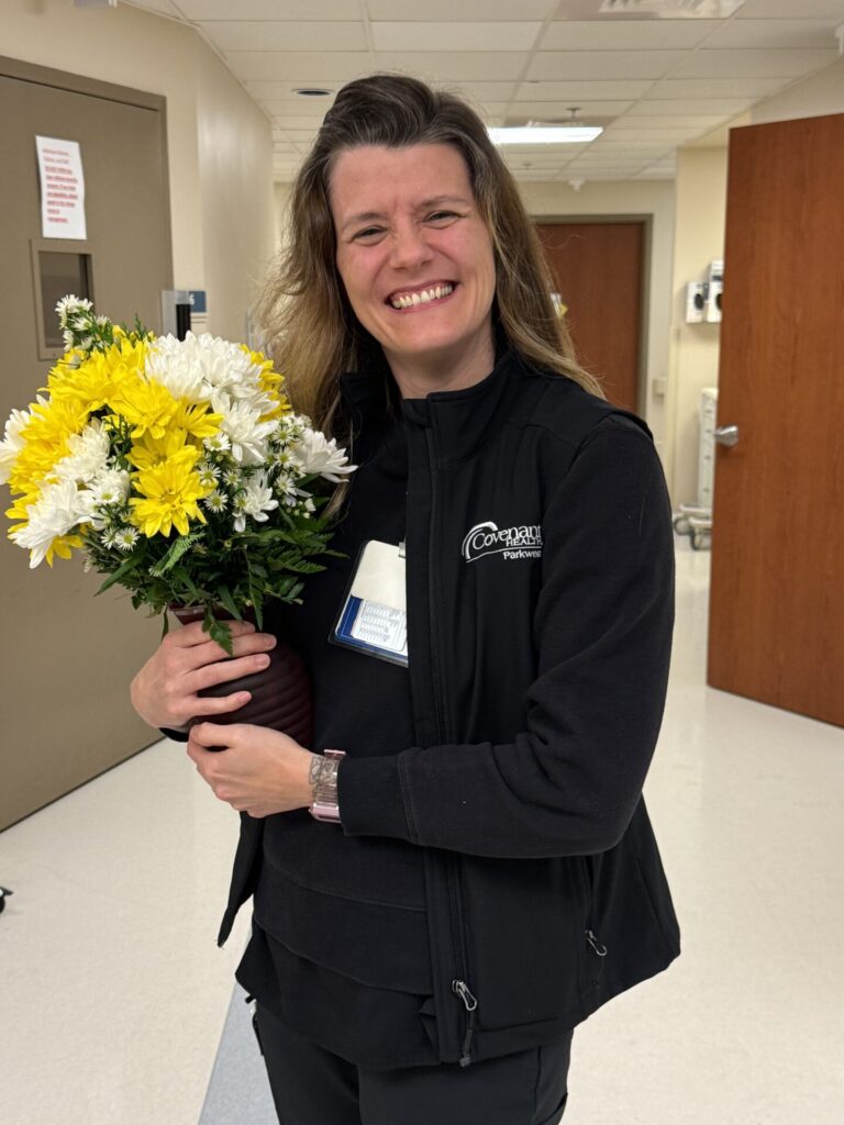 A woman smiles, holding a bouquet of yellow and white flowers in a hallway with beige walls and wooden doors, wearing a black jacket with a work badge&mdash;celebrating as Ethan Howe wins the DAISY Award.
