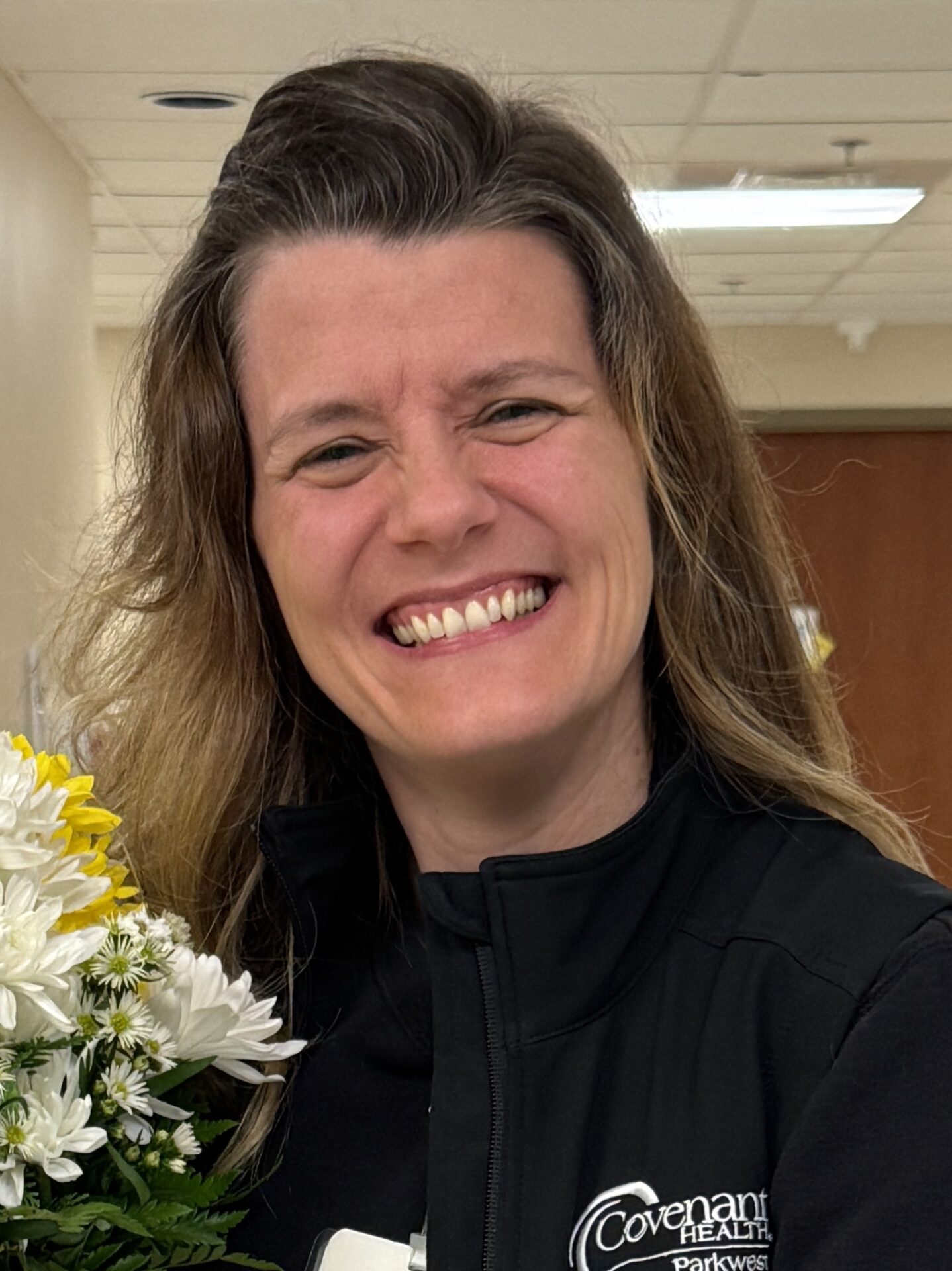 A woman with long brown hair smiles brightly while holding a bouquet of white and yellow flowers, celebrating as Ethan Howe wins DAISY Award. She wears a black jacket with the Covenant Health Parkwest logo in a hallway with beige walls.