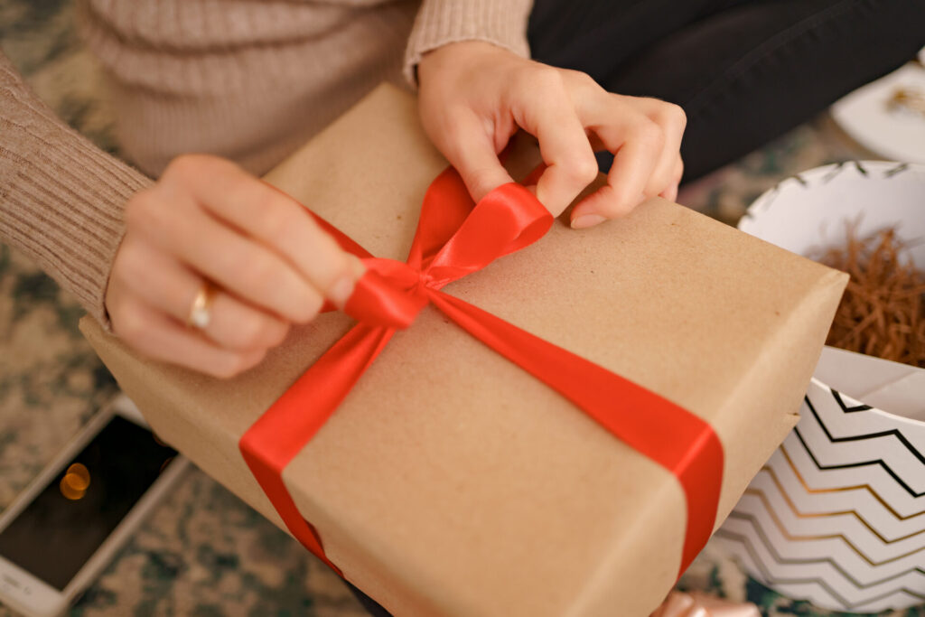 A person ties a red satin ribbon on a gift wrapped in brown paper.