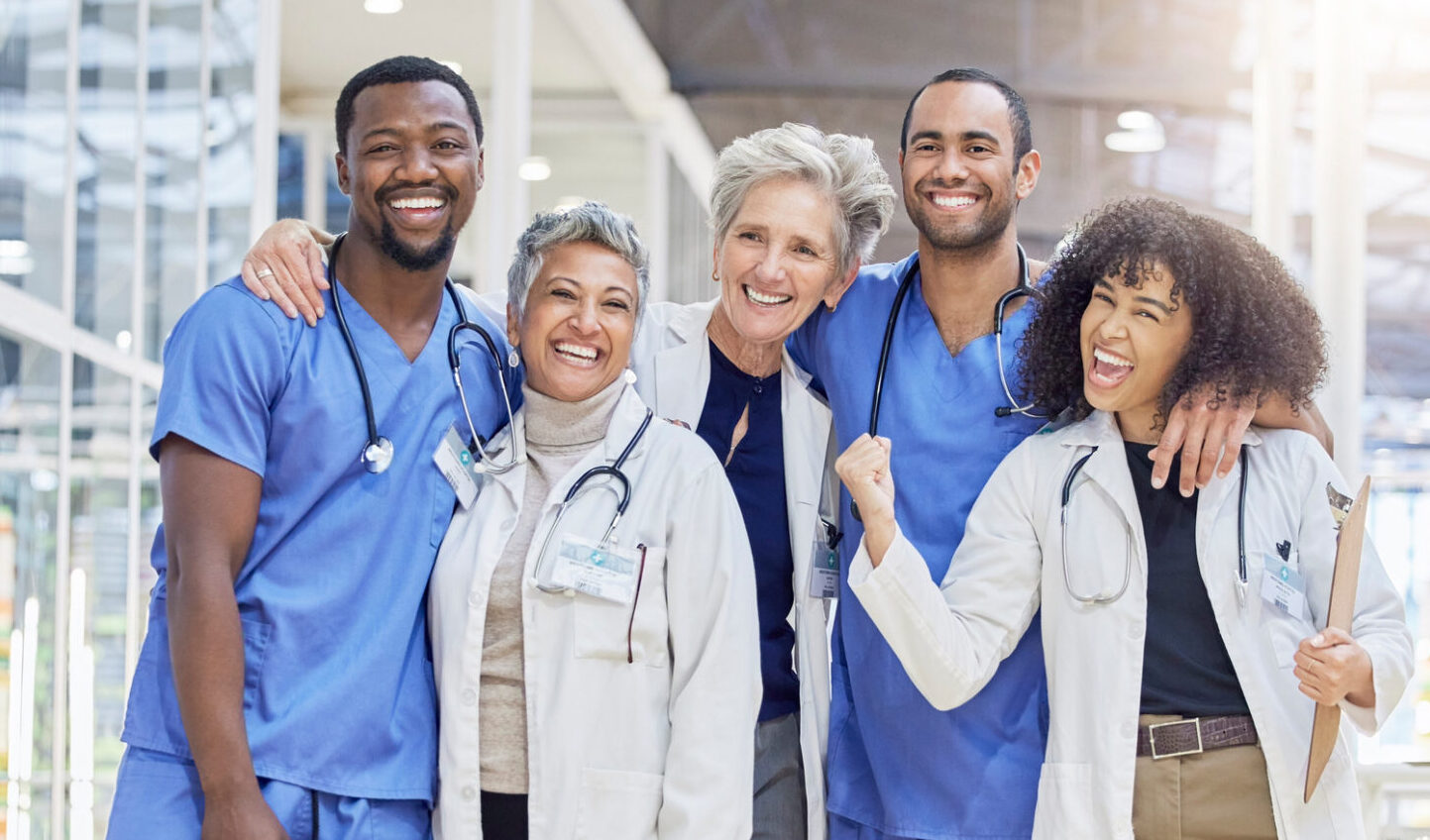 A diverse group of five smiling healthcare professionals, wearing scrubs and white coats, stand together in a bright, modern hospital corridor, with arms around each other, showing camaraderie and teamwork.