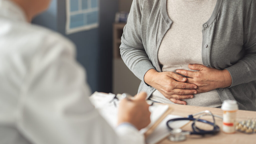 A person sits at a doctors office holding their abdomen as if in pain while speaking with a healthcare professional; a stethoscope, medication bottle, and clipboard are on the desk.