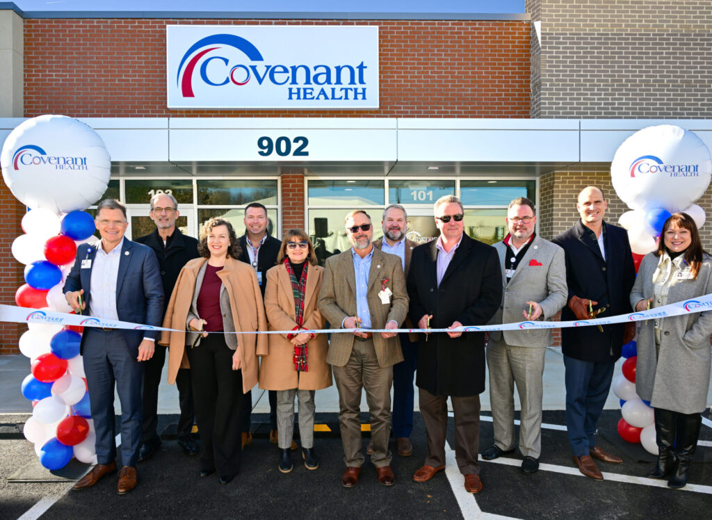 A group of people stands in front of a Covenant Health building, smiling and holding a ribbon for a grand opening. Balloon columns with red, white, and blue balloons flank the entrance.