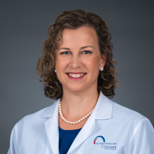 A smiling woman with curly light brown hair, wearing a white medical coat, blue shirt, pearl necklace, and pearl earrings, poses in front of a plain gray background.