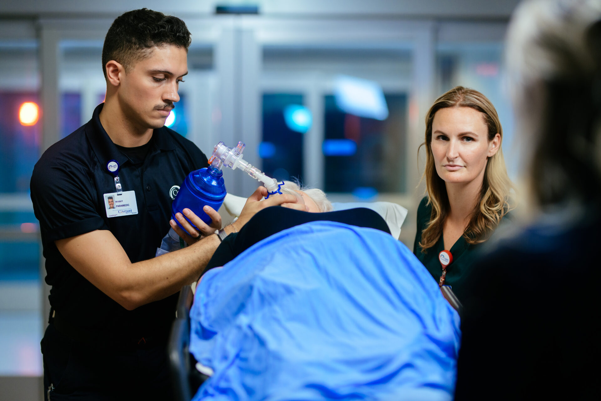 A medical professional uses a manual resuscitator on a patient lying on a stretcher, while another healthcare worker stands nearby, looking attentively. The scene is in a brightly lit hospital setting.