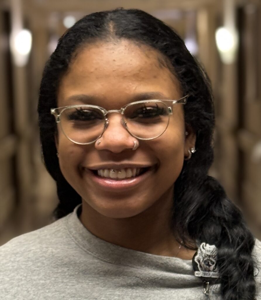 A person with long dark hair, glasses, and a nose ring smiles at the camera. They wear a gray top with a pin featuring a wolf logo and an Ahniah Ray BEE Award. The softly blurred background resembles a hallway with lights.