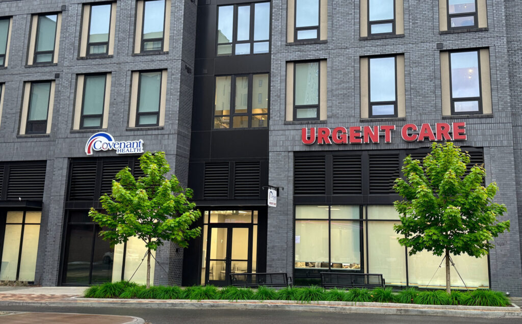 A modern gray brick building with large windows houses two businesses: Covenant Eye Care on the left and a bold red Urgent Care sign on the right. Three green trees line the sidewalk in front.