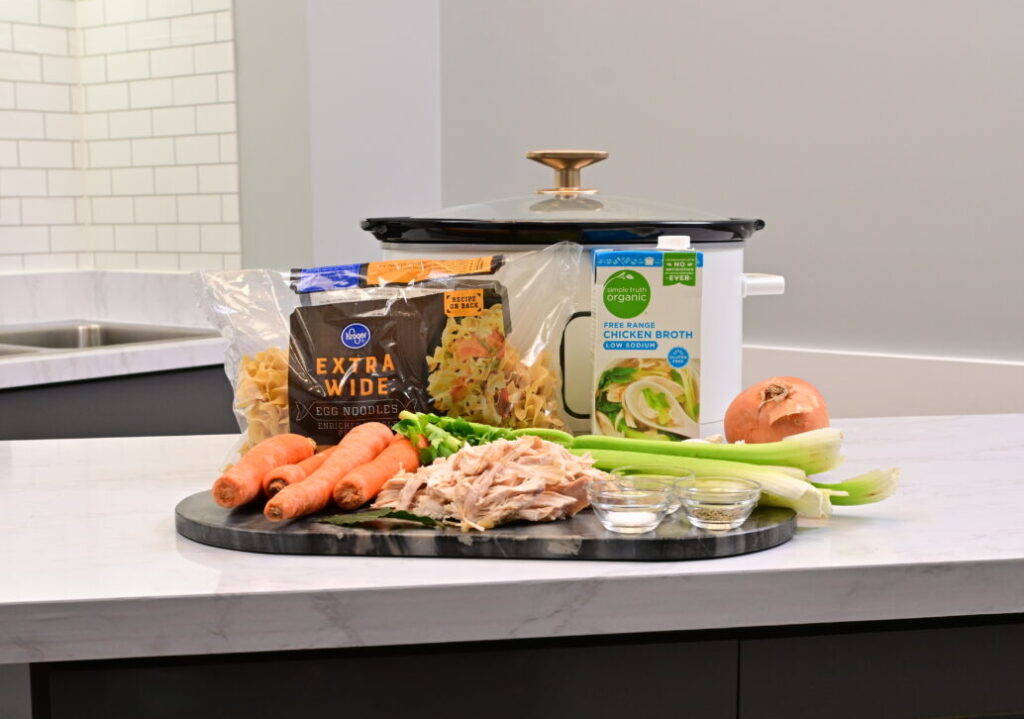 A crockpot sitting on a countertop surrounded by the ingredients for chicken noodle soup