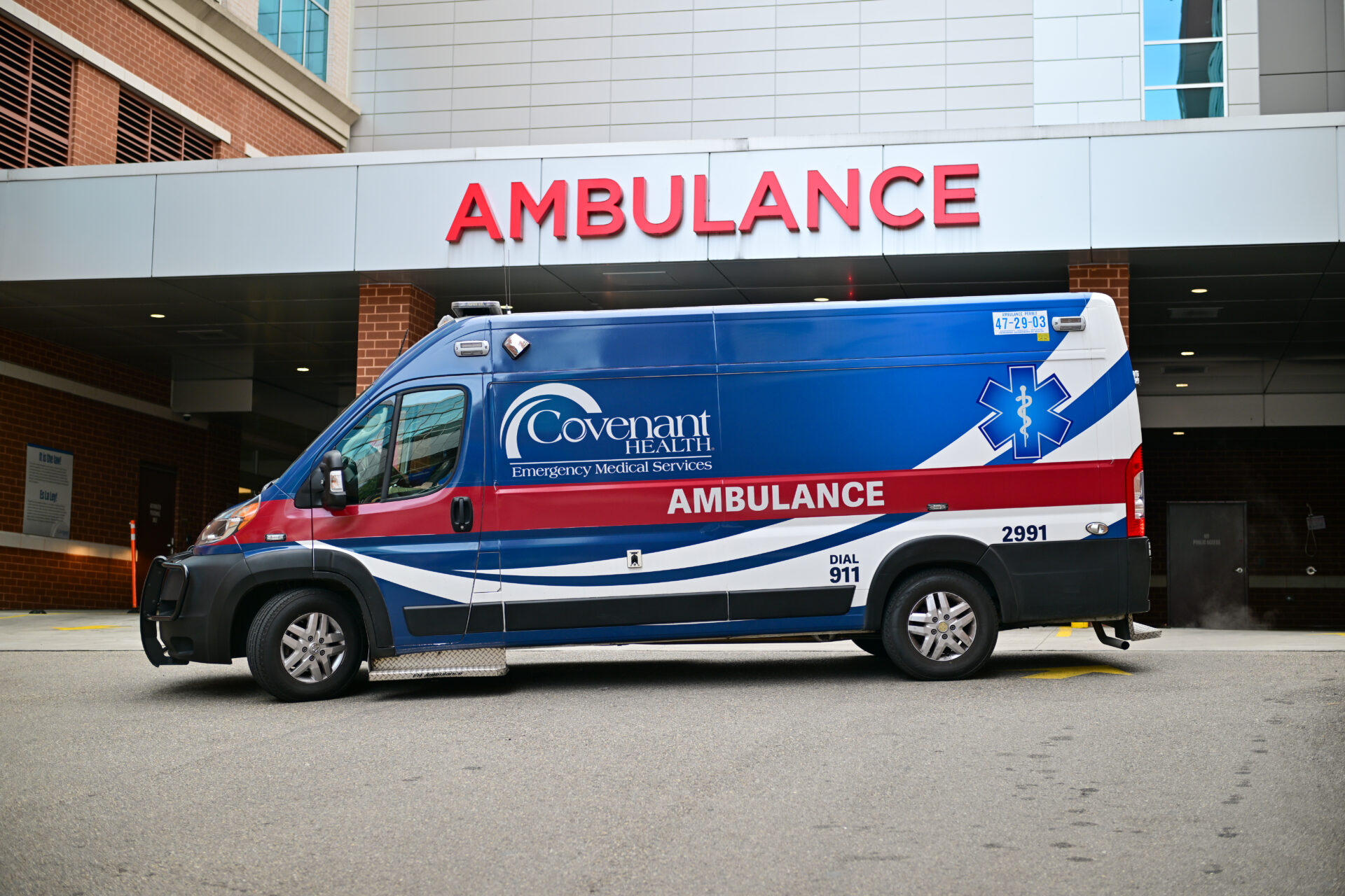 A blue and white Covenant Health ambulance is parked outside a hospital emergency entrance with a large red AMBULANCE sign above the doors.