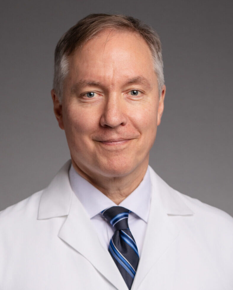 A middle-aged man with short gray hair, wearing a white lab coat and blue striped tie, stands against a plain gray background, smiling slightly and facing the camera—one of the dedicated Covenant Health Proton Center Physicians.