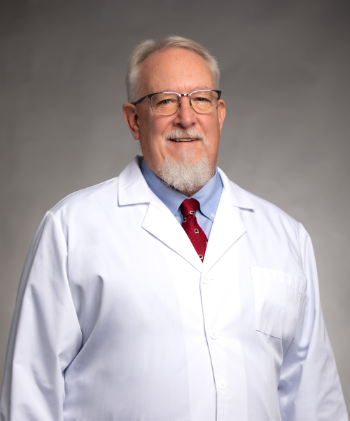 An older man with white hair, glasses, and a beard is wearing a white lab coat, blue shirt, and red patterned tie, standing against a neutral gray background.