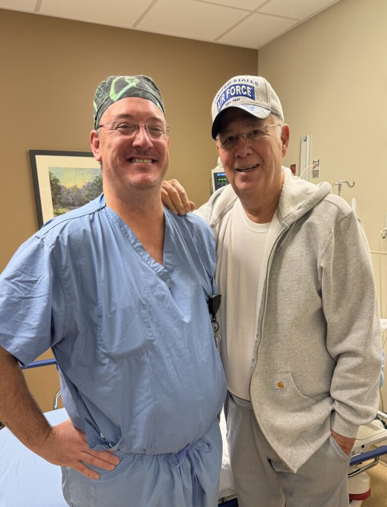 Two men, Dr. Copertino and Gerald Smith are smiling standing in a hospital room. One is wearing blue surgical scrubs and a cap; the other is dressed casually in a gray hoodie and cap that says Air Force. They are standing close together, appearing cheerful.