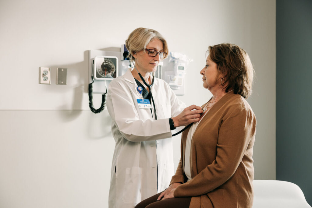 A doctor in a white coat uses a stethoscope to listen to a seated womans chest during a medical exam in a clinic room. Medical equipment is mounted on the wall in the background.