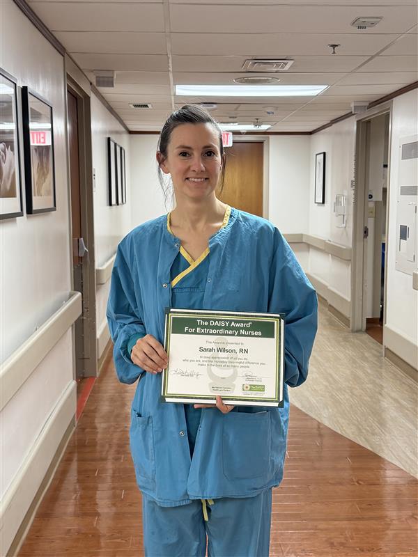 A nurse in blue scrubs stands in a hospital hallway, smiling and holding a certificate that reads The DAISY Award for Extraordinary Nurses. The hallway has wooden floors and framed pictures on the walls.