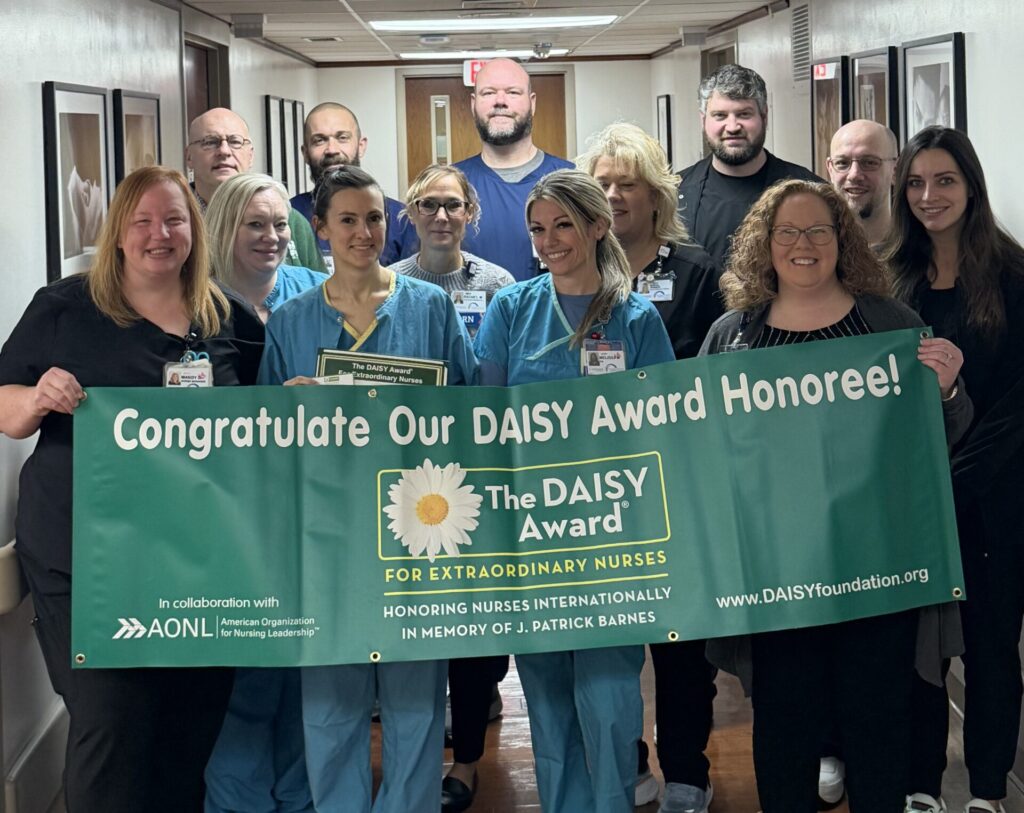 A group of hospital staff, including nurses in scrubs and others in professional attire, stand in a hallway holding a green banner congratulating a DAISY Award honoree for extraordinary nurses.