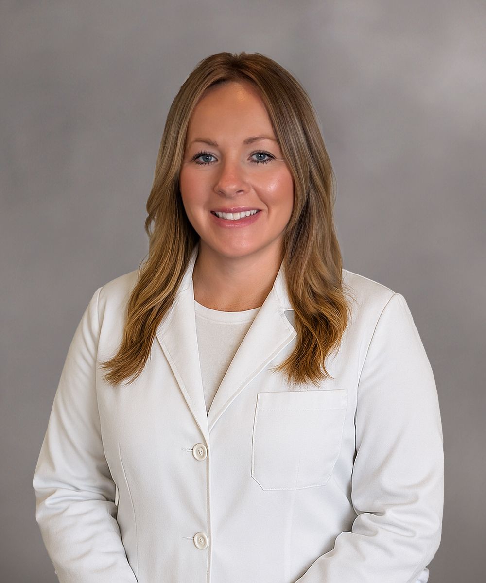 A woman with long, light brown hair is smiling and wearing a white lab coat over a light top. She is standing in front of a plain, gray background.