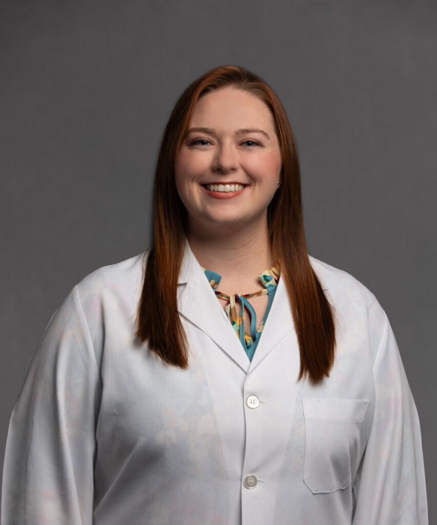 A woman with long, straight reddish-brown hair smiles and stands facing the camera. She wears a white lab coat over a patterned blouse. The background is plain and gray.