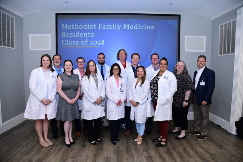 A group of medical professionals in white coats and business attire stands in front of a screen reading Methodist Family Medicine Residents Class of 2028, posing and smiling for a group photo in a well-lit room.