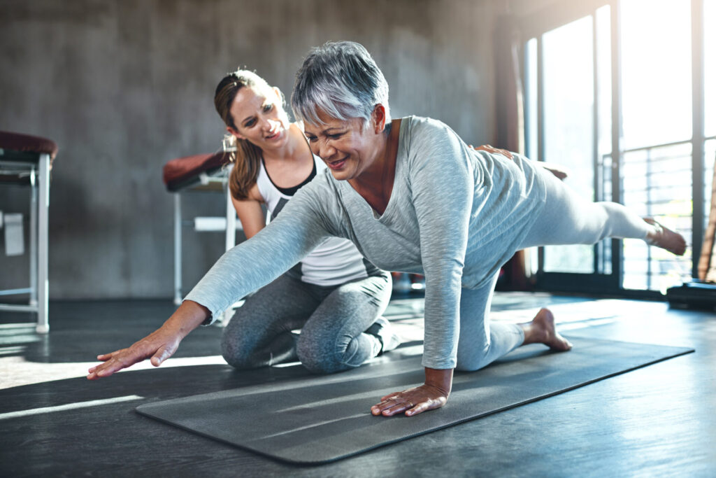 An older woman in workout clothes balances on one knee and hand while extending her opposite arm and leg. A younger woman kneels beside her, offering guidance and encouragement in a bright fitness studio.
